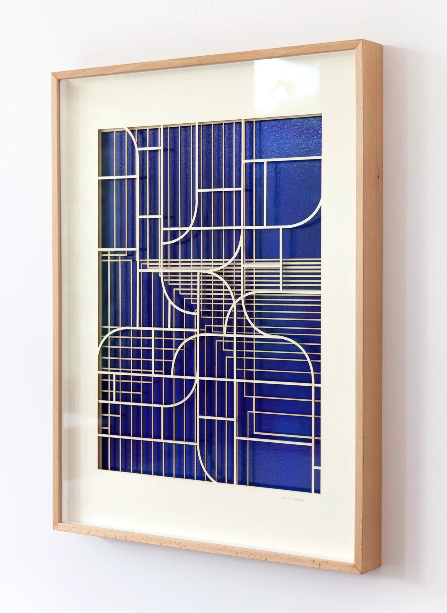 Oblique angled view of geometric abstract artwork by Jesús Perea, showing the overlapping laser-cut museum board layers that create a three-dimensional relief over the blue background.