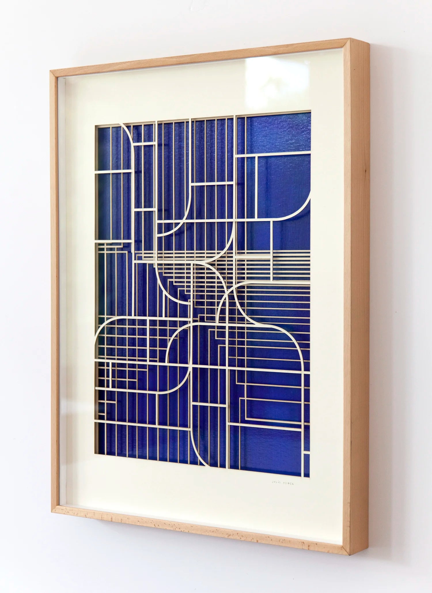 Oblique angled view of geometric abstract artwork by Jesús Perea, showing the overlapping laser-cut museum board layers that create a three-dimensional relief over the blue background.